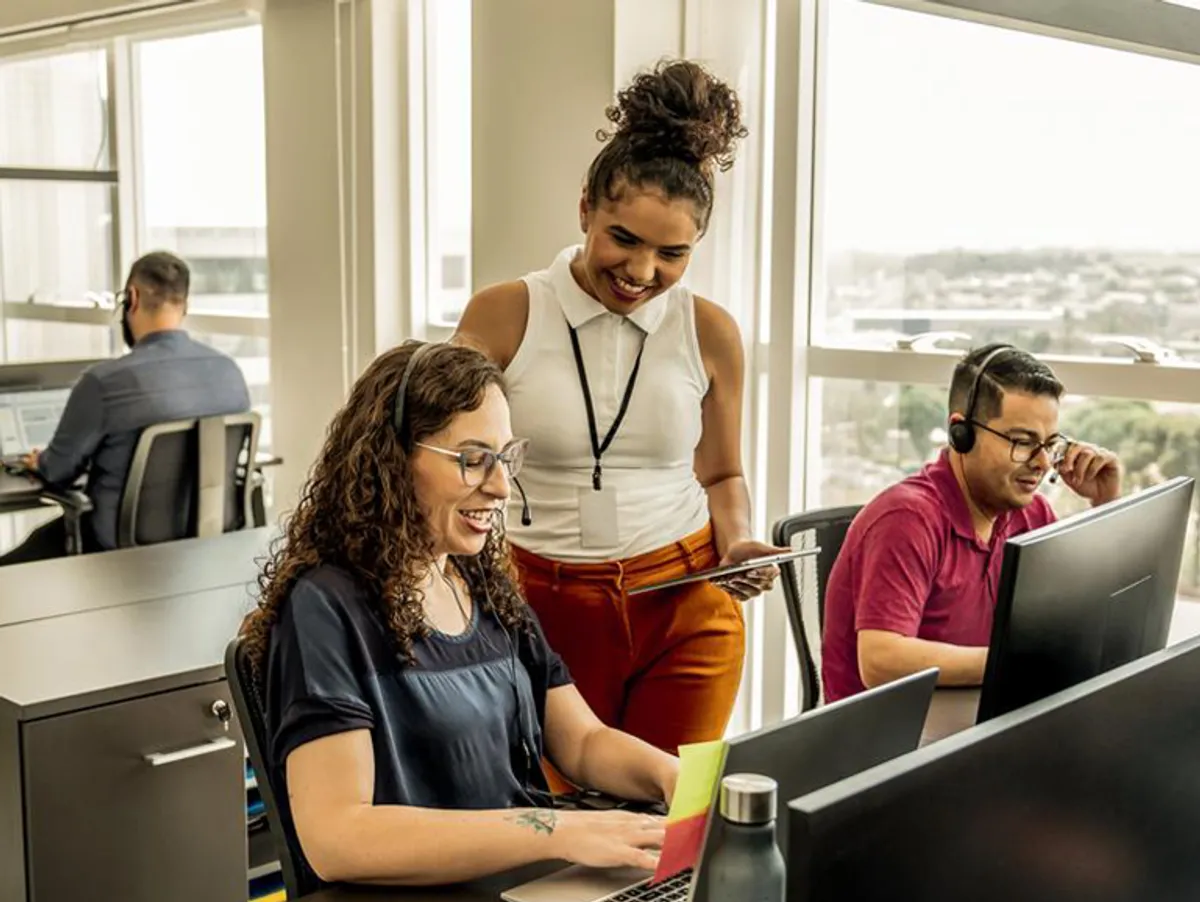 Three colleagues in an office working at desks with laptops, wearing headsets, and conversing with each other.